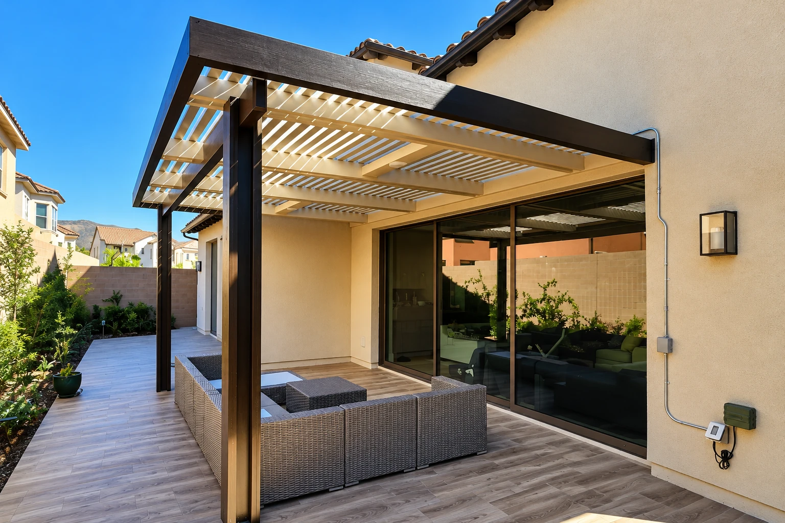 A dark wood patio cover with slatted panels shades a wicker seating area beside a stucco house and sliding glass doors.