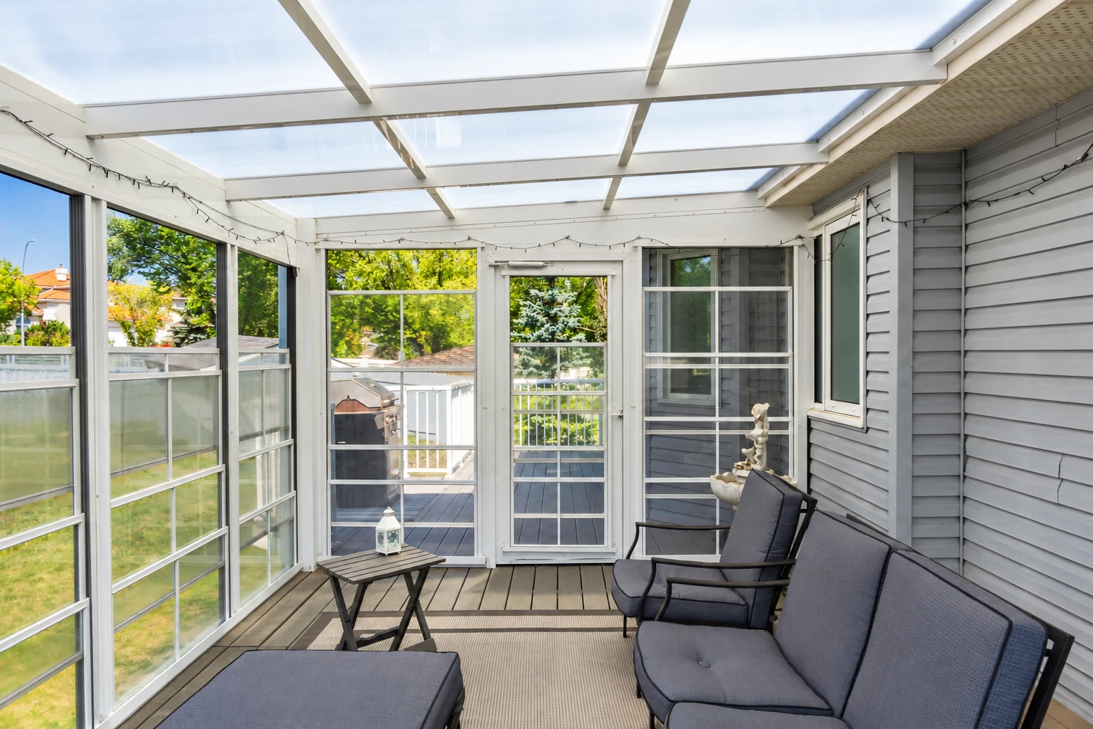 Enclosed patio with a translucent roof, white-framed windows, gray siding, and cushioned lounge chairs.