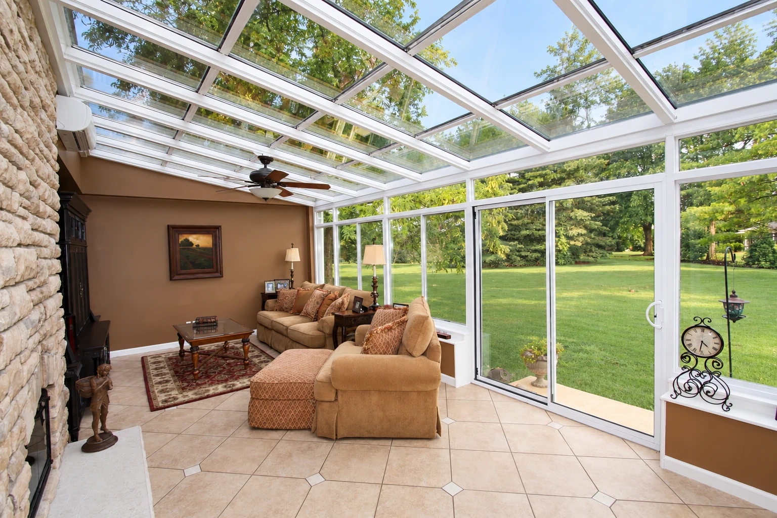 A solarium with a glass-paneled roof and white-framed windows has tiled floors, tan seating, and views of a green lawn.