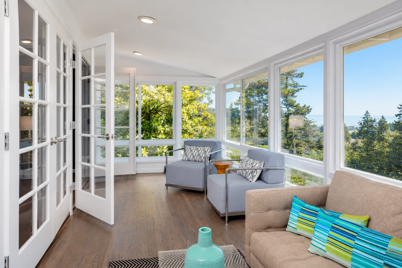 Sunroom with white-framed windows and open French doors, furnished with pale blue chairs and a beige sofa.