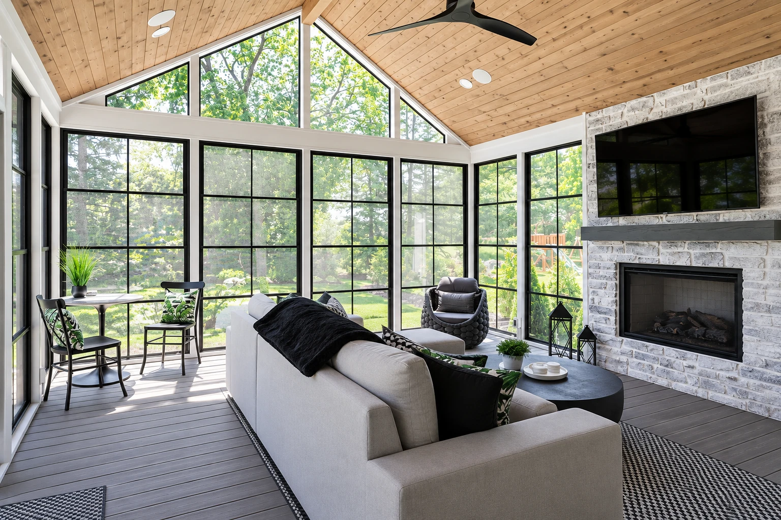 A bright three-season sunroom with a wood-plank vaulted ceiling, black-grid windows, a stone fireplace, and neutral lounge seating.