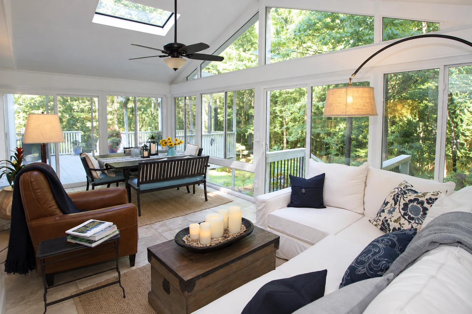 A white-framed vinyl sunroom with expansive windows, a skylight, a dining table, and white sofas overlooking trees.